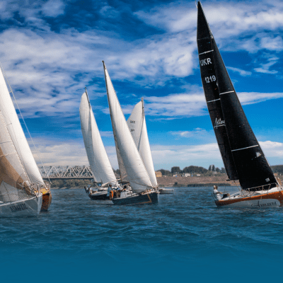 Several sailboats racing on a river under a cloudy sky with a bridge in the background.